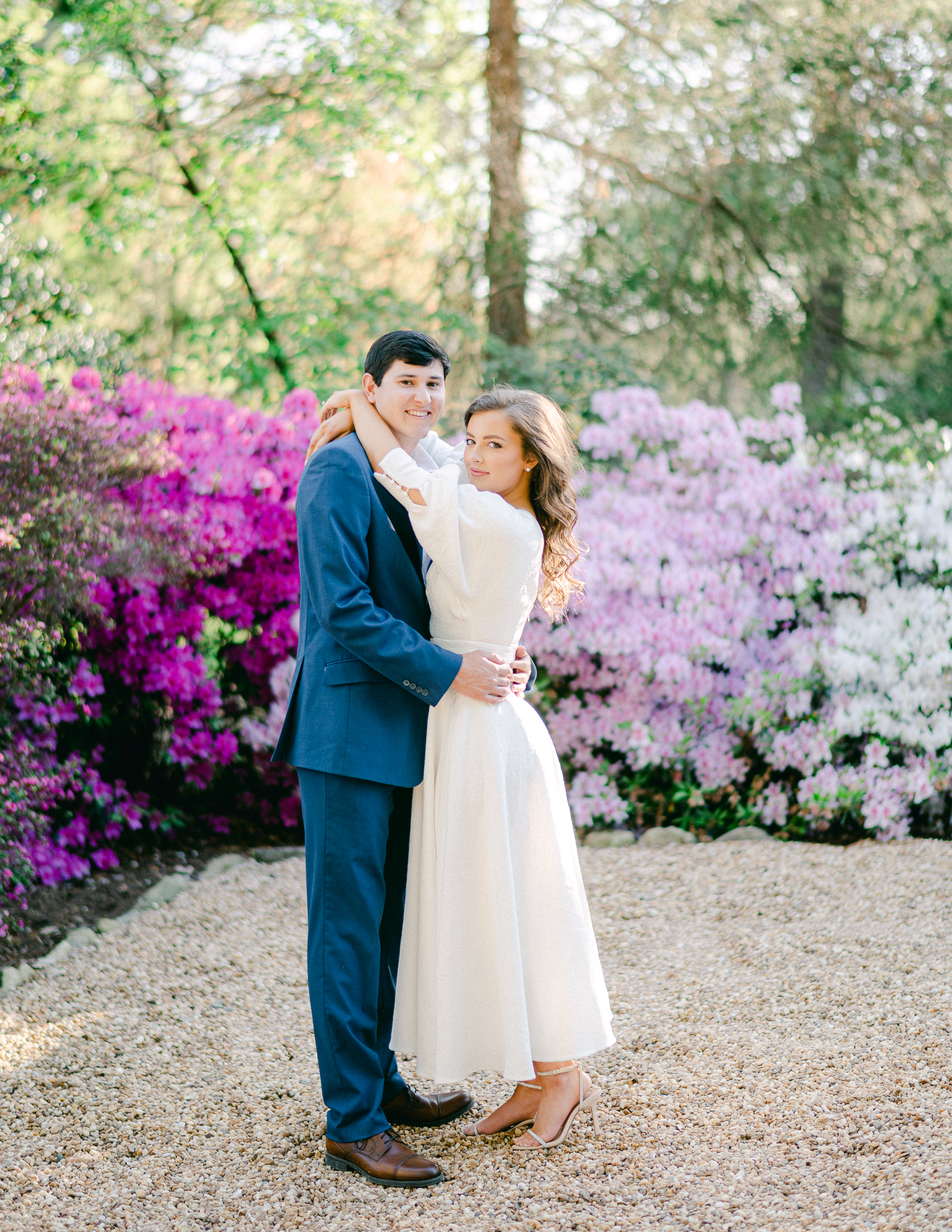 girl in a long white dress with guy in a navy blue suit with a bed of pink & purple florals behind them.